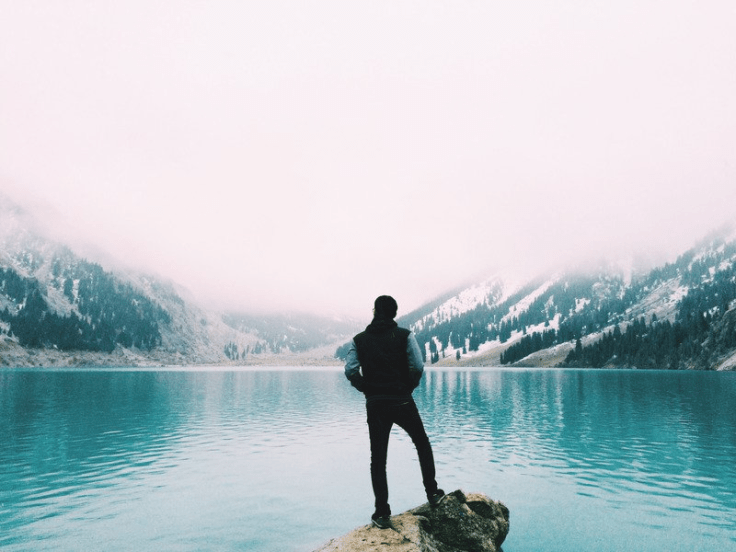 Man in solitude looking at a glacial lake surrounded by snow and pine clad mountains